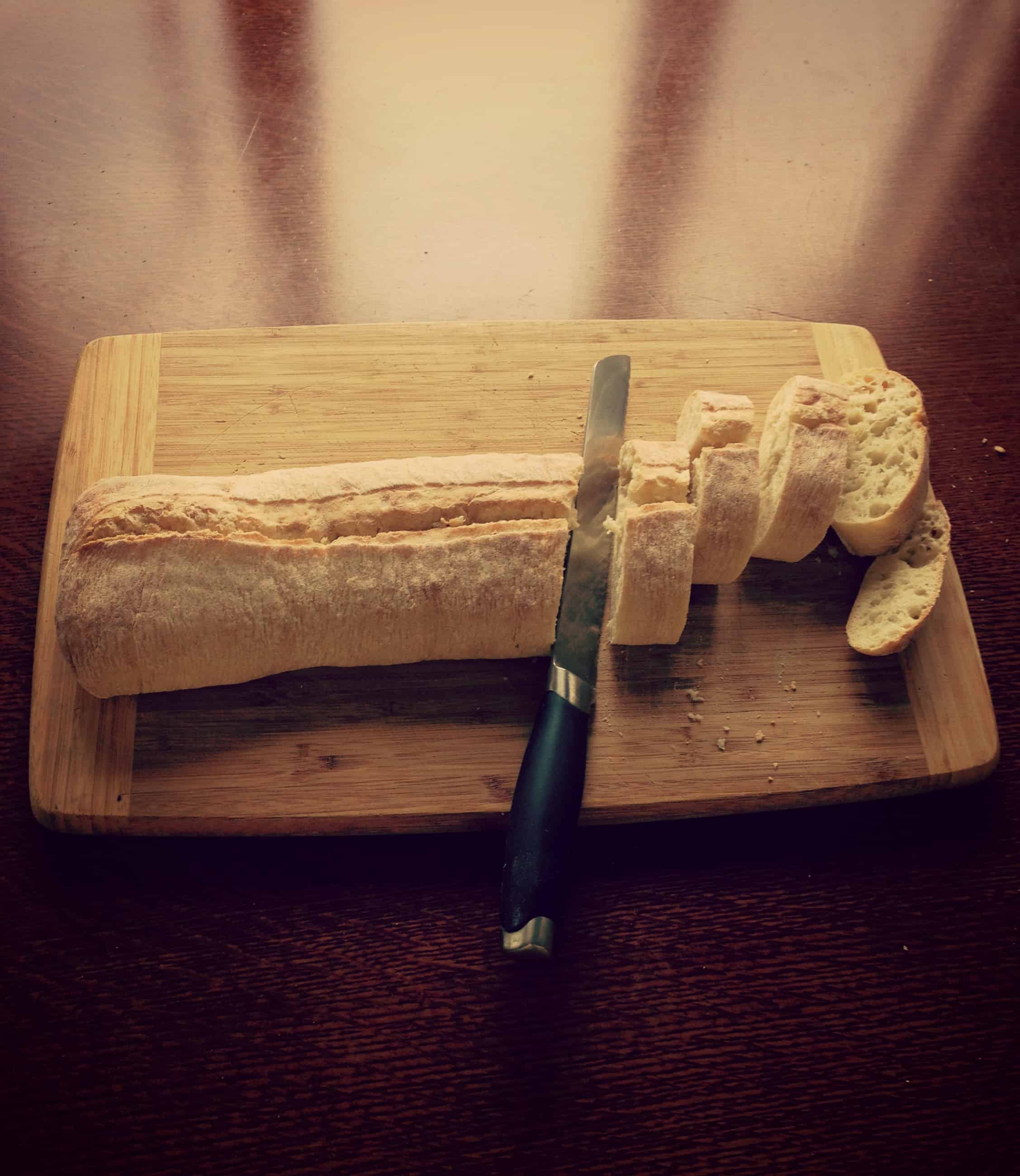 A bread knife and loaf of half sliced baguette on a wood cutting board.