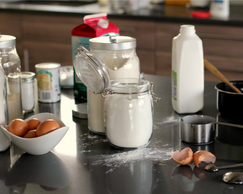 Jars of flour and sugar with a milk carton and a bowl of eggs.