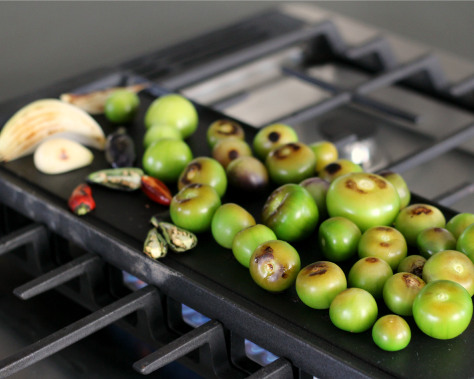 Tomatillos and chili peppers roasting on a griddle.