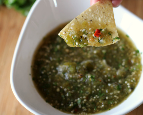 A tortilla chip holding a scoop of salsa verde from a white bowl.