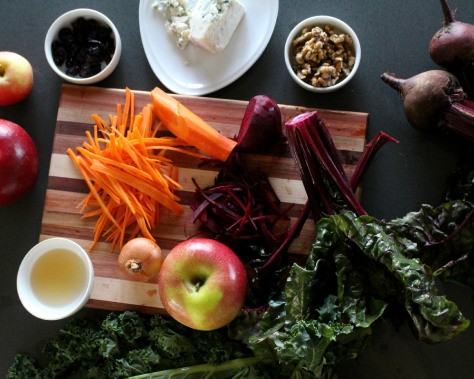 A wood cutting board with kale, apples, carrots, and onions.