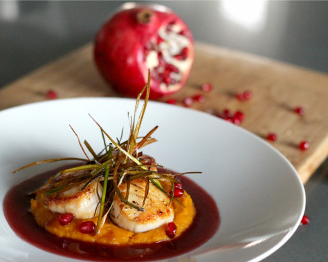 Seared scallops in a shallow white bowl with pomegranate arils and fresh herbs.