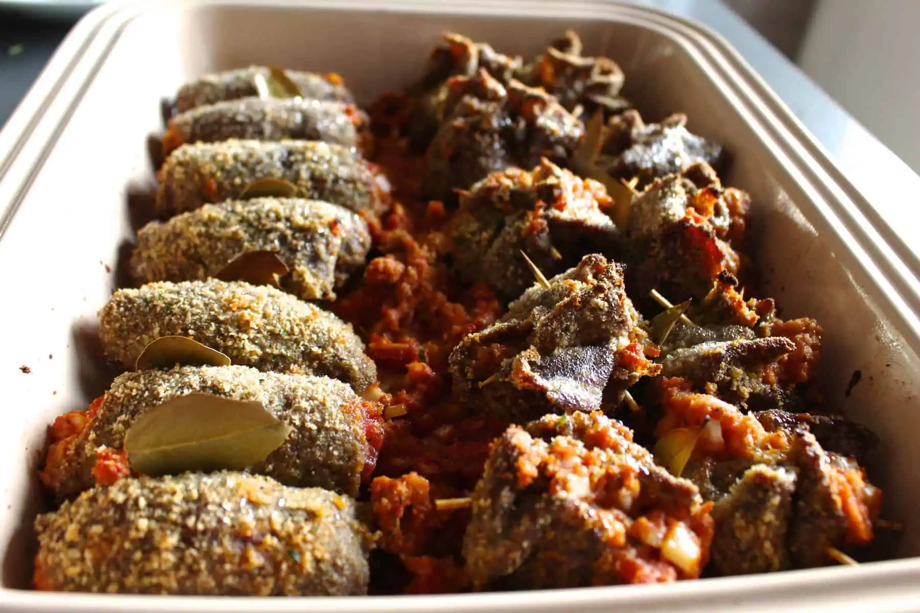 Thinly sliced beef wrapped around stuffed tomatoes in a baking dish.