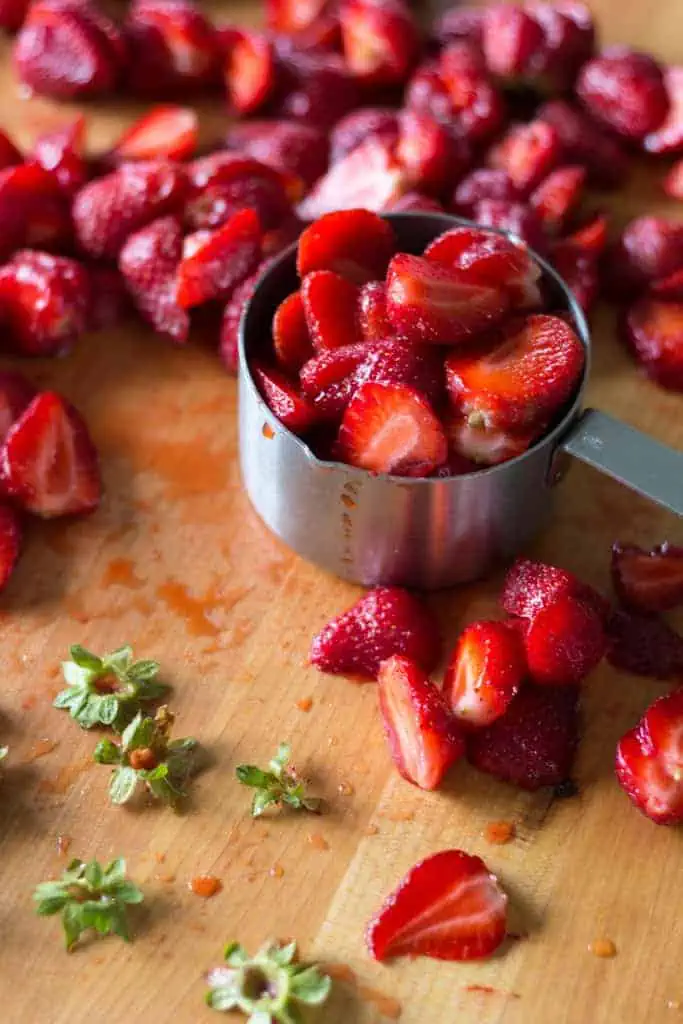A metal measuring cup filled with strawberry halves on a wood cutting board surrounded by strawberries.