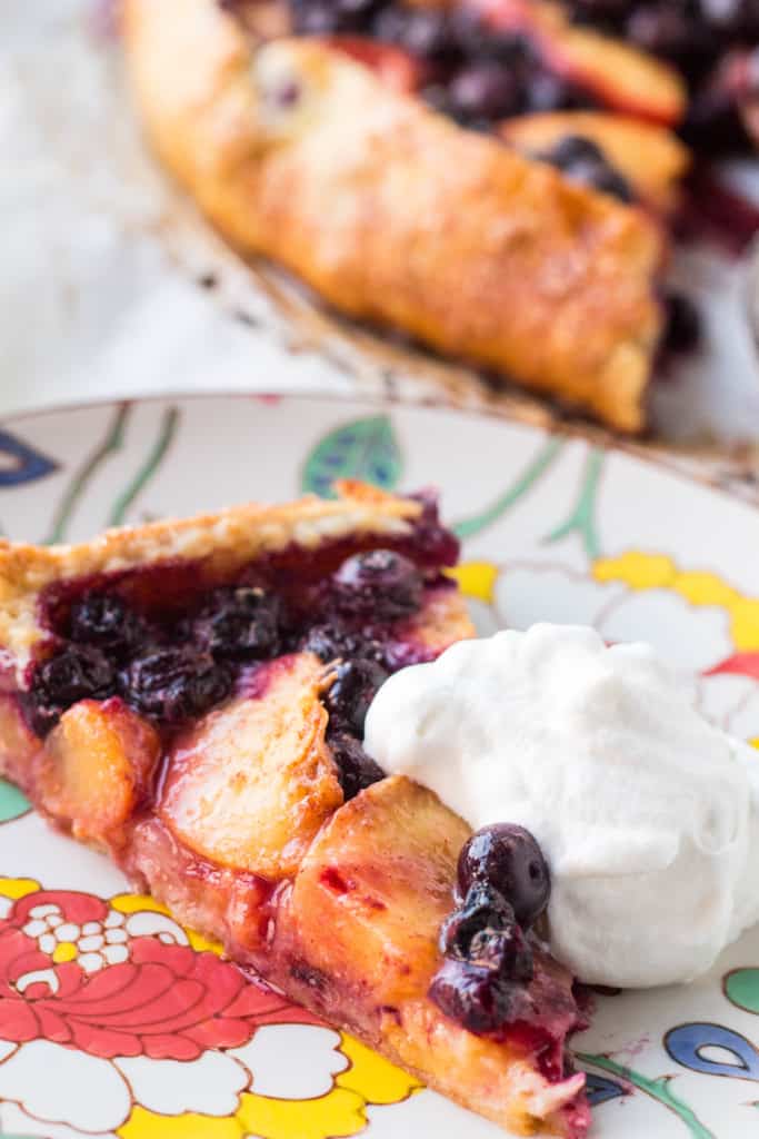 Close up of a slice of blueberry peach crostata and a dollop of whipped cream on a plate.