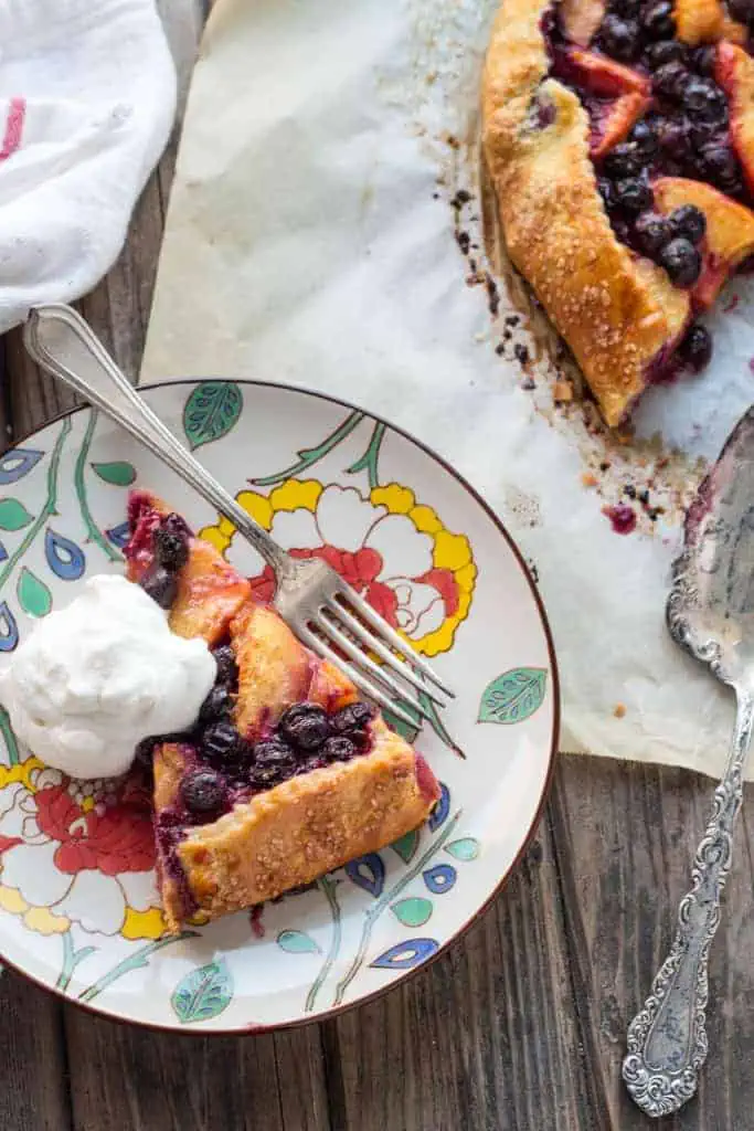 Top view of a floral plate holding a fork, slice of blueberry peach crostata and a dollop of whipped cream.