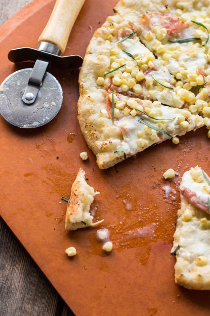 Top view of slices of grilled corn and prosciutto pizza next to a pizza cutter a bite of crust.