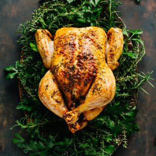 whole roasted chicken on a serving platter surrounded by fresh herbs on dark wood background