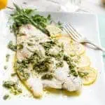 Close up of an herb roasted barramundi fillet next to lemon rounds and a fork on a white serving dish.