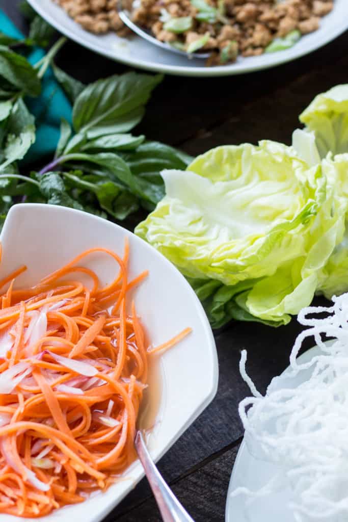 Close up of a bowl of shredded, pickled carrots next to butter lettuce leaves.