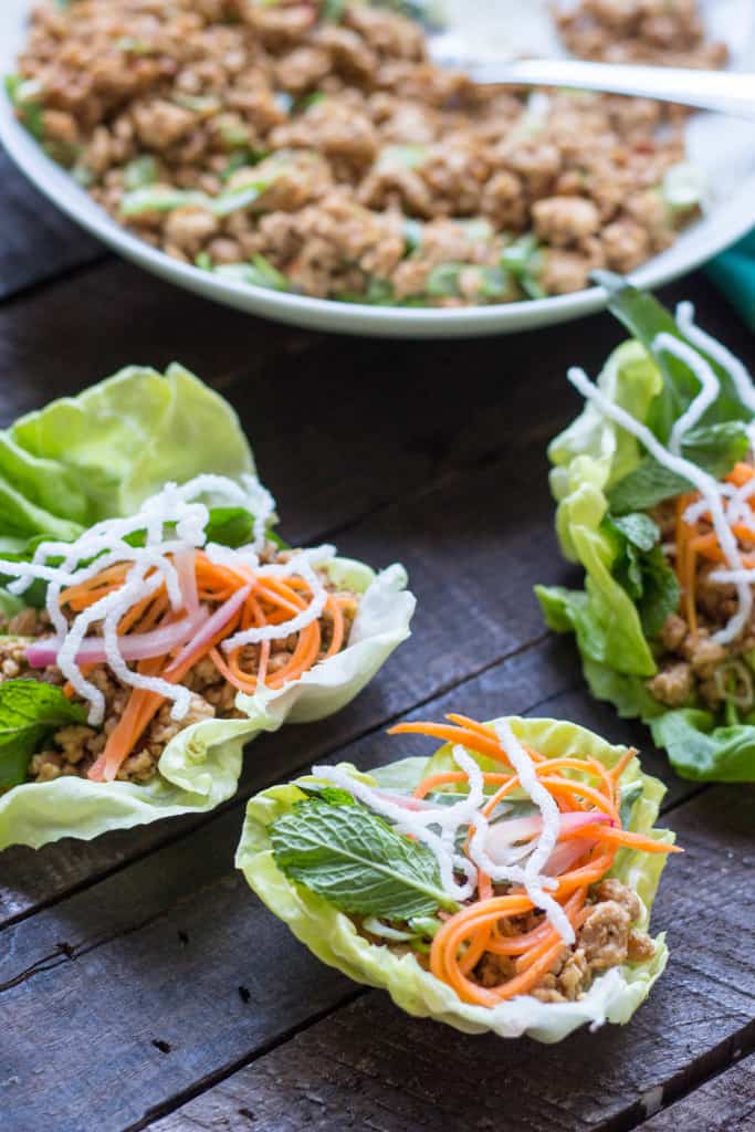 Close up of three lettuce wraps filled with chicken, carrots, and rice noodles on a wood table.