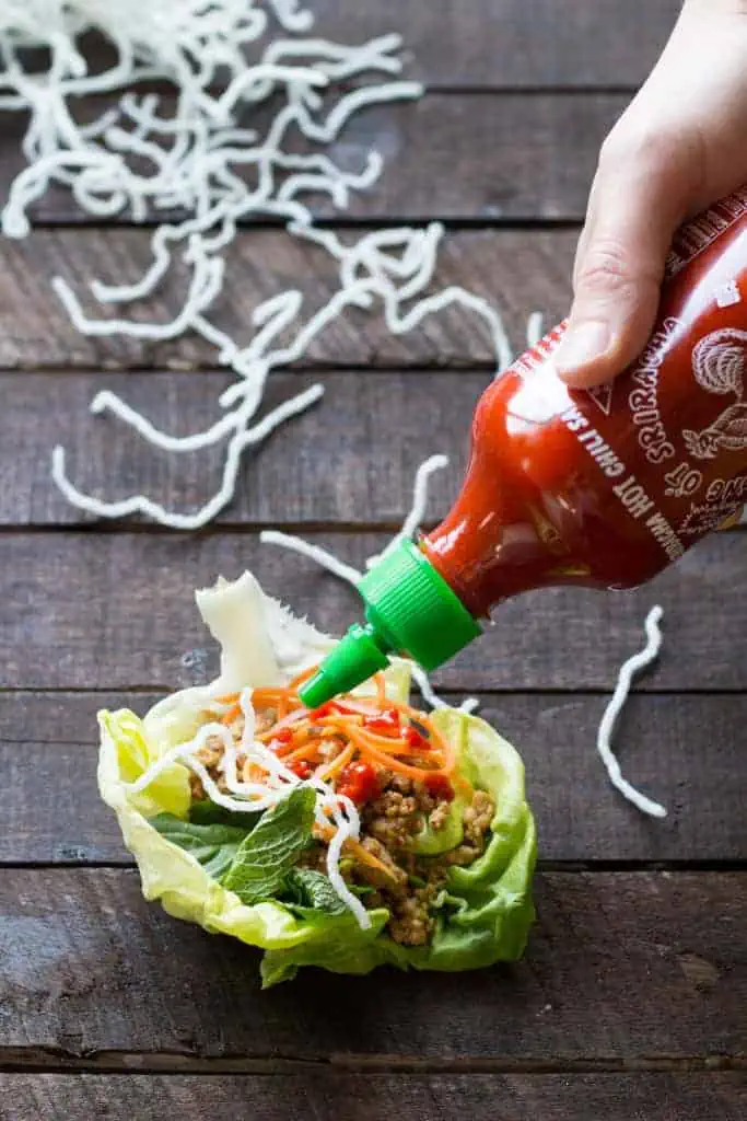 A hand holding a bottle of sriracha over a chicken lettuce cup.