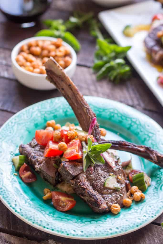 Close up of an aqua dinner plate with two lamb chops topped with tomato cucumber salad and crispy chickpeas.