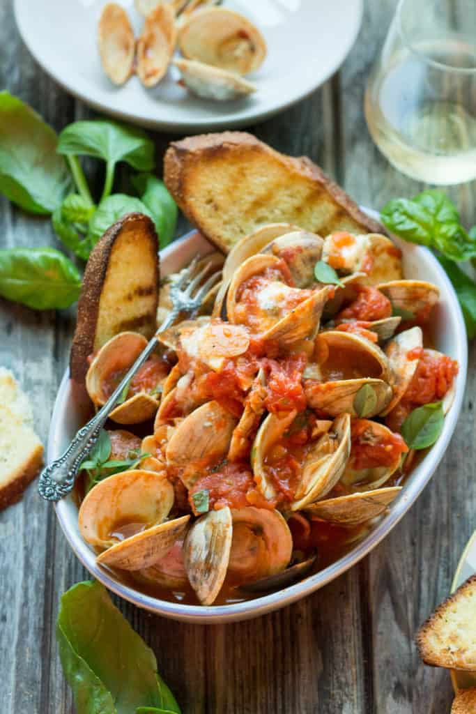 White serving bowl of steamed clams with toasted baguette slices.