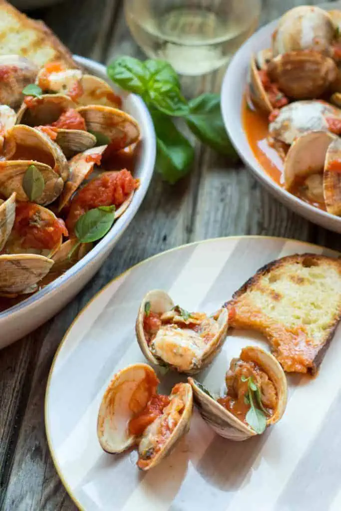 Three tomato steamed clams on a white dinner plate with a half eaten slice of toasted baguette.