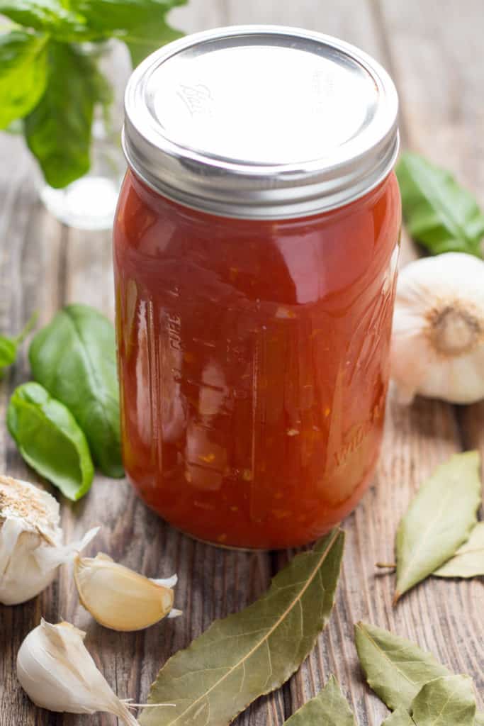 Quart jar of homemade Italian red gravy surrounded by basil leaves, bay leaves, and garlic heads on a wood board.