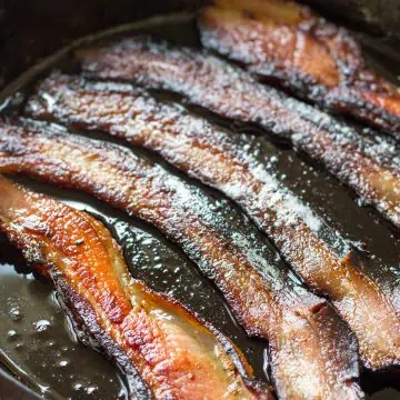 Five slices of bacon frying in a cast iron skillet.