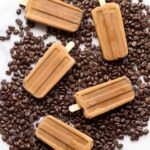 Homemade creamy coffee popsicles arranged on a bed of roasted coffee beans over a marble surface, showcasing their rich mocha color and smooth texture.