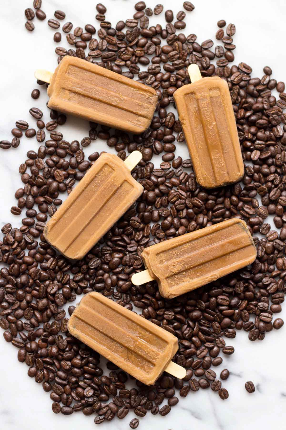 Homemade creamy coffee popsicles arranged on a bed of roasted coffee beans over a marble surface, showcasing their rich mocha color and smooth texture.