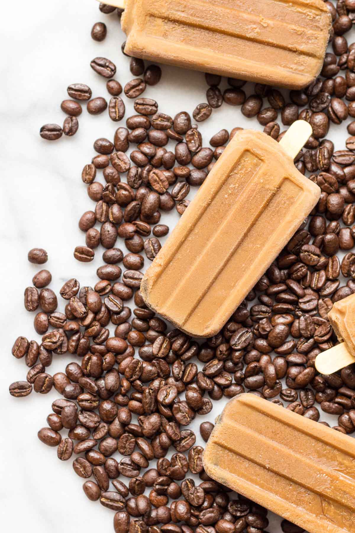 Close-up of frozen coffee popsicles resting on whole roasted coffee beans, highlighting the icy details and velvety brown hue of these refreshing summer treats.