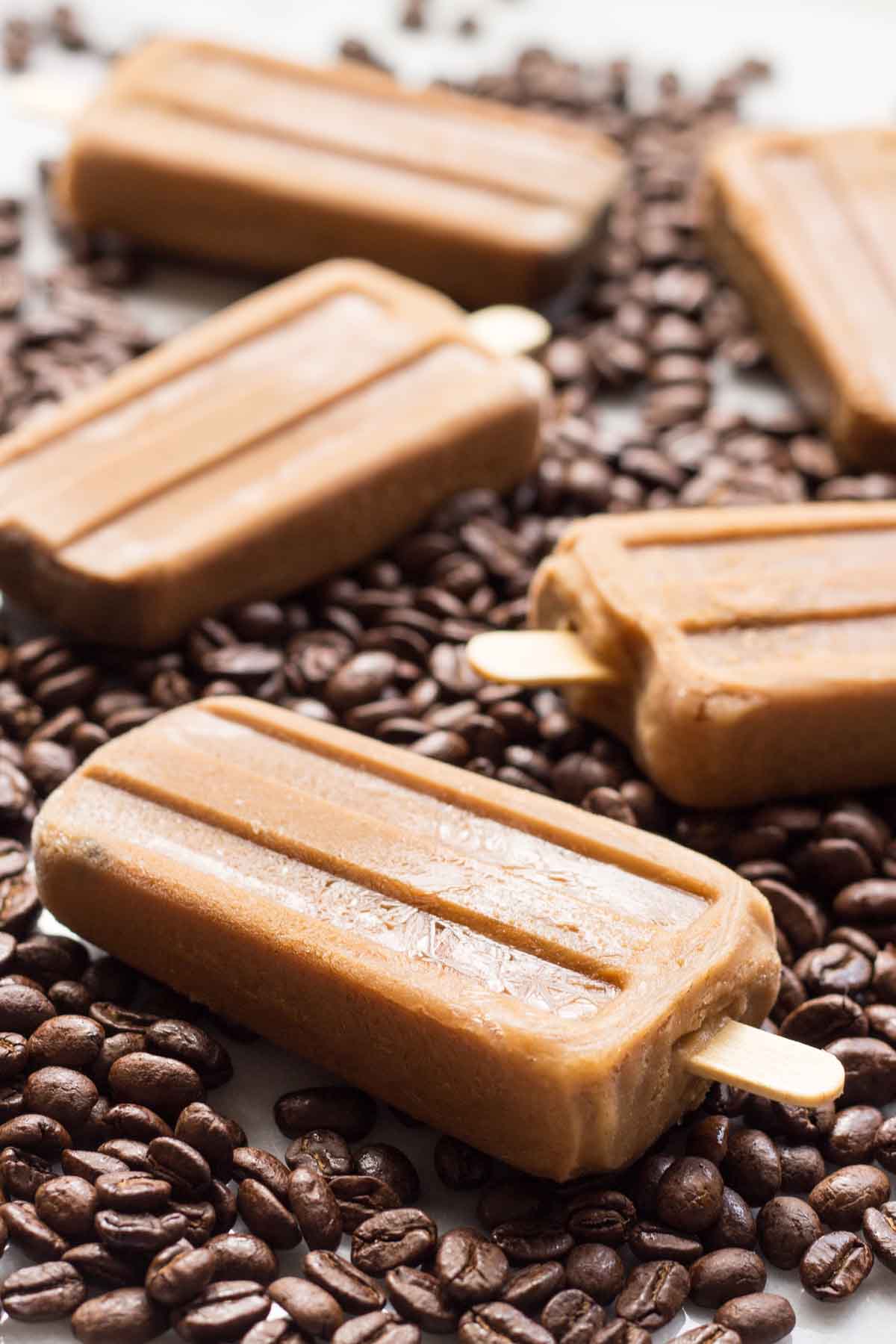 Angled side view of frozen coffee popsicles on a backdrop of coffee beans, emphasizing the popsicles’ creamy texture and bold espresso color.