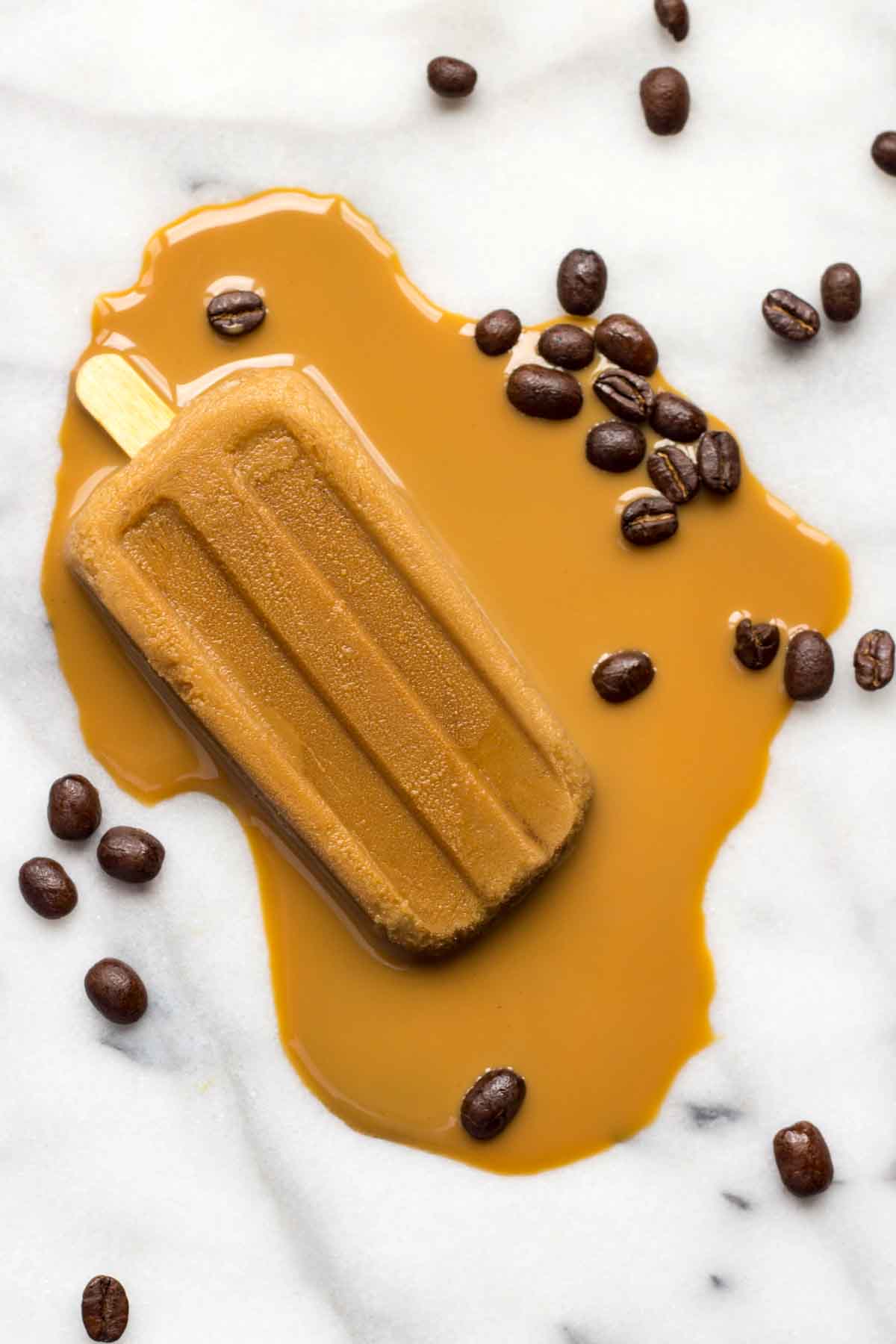 Melted coffee popsicle on a white marble background surrounded by scattered roasted coffee beans, with coffee pooling dramatically around the base—perfect for a summer dessert concept.