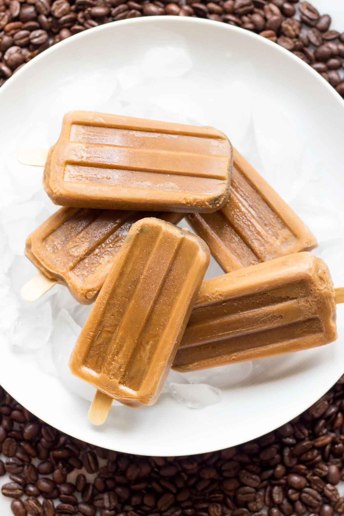 Coffee popsicles stacked in a white bowl filled with ice, placed on a ring of coffee beans—showing a refreshing and caffeinated frozen treat ideal for hot days.