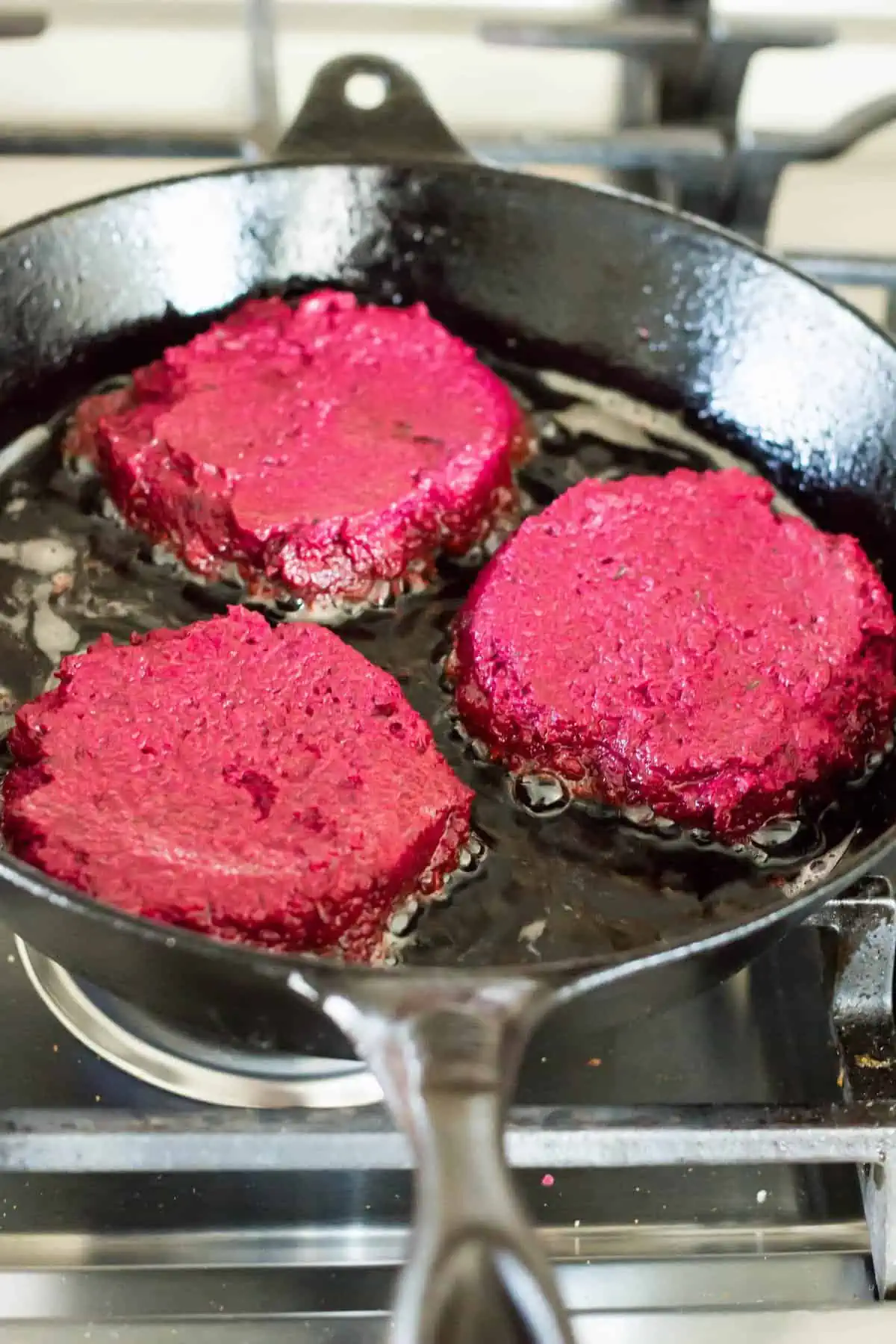 Three beautiful pink lentil beet burgers frying in a cast iron skillet.