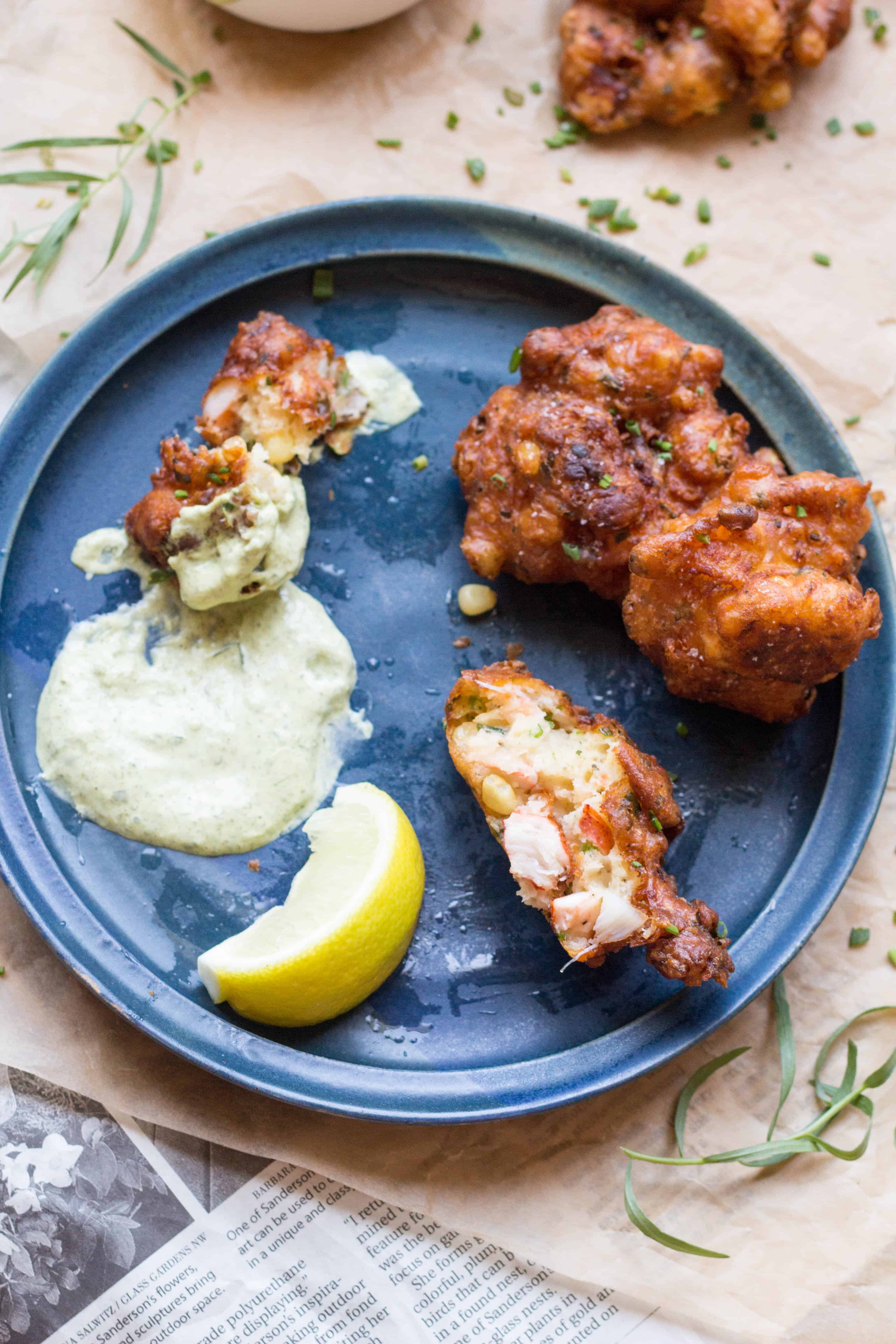 Top view of a blue plate with a lemon wedge, two corn fritters, and one fritter dipped into a pool of tarragon aoli.