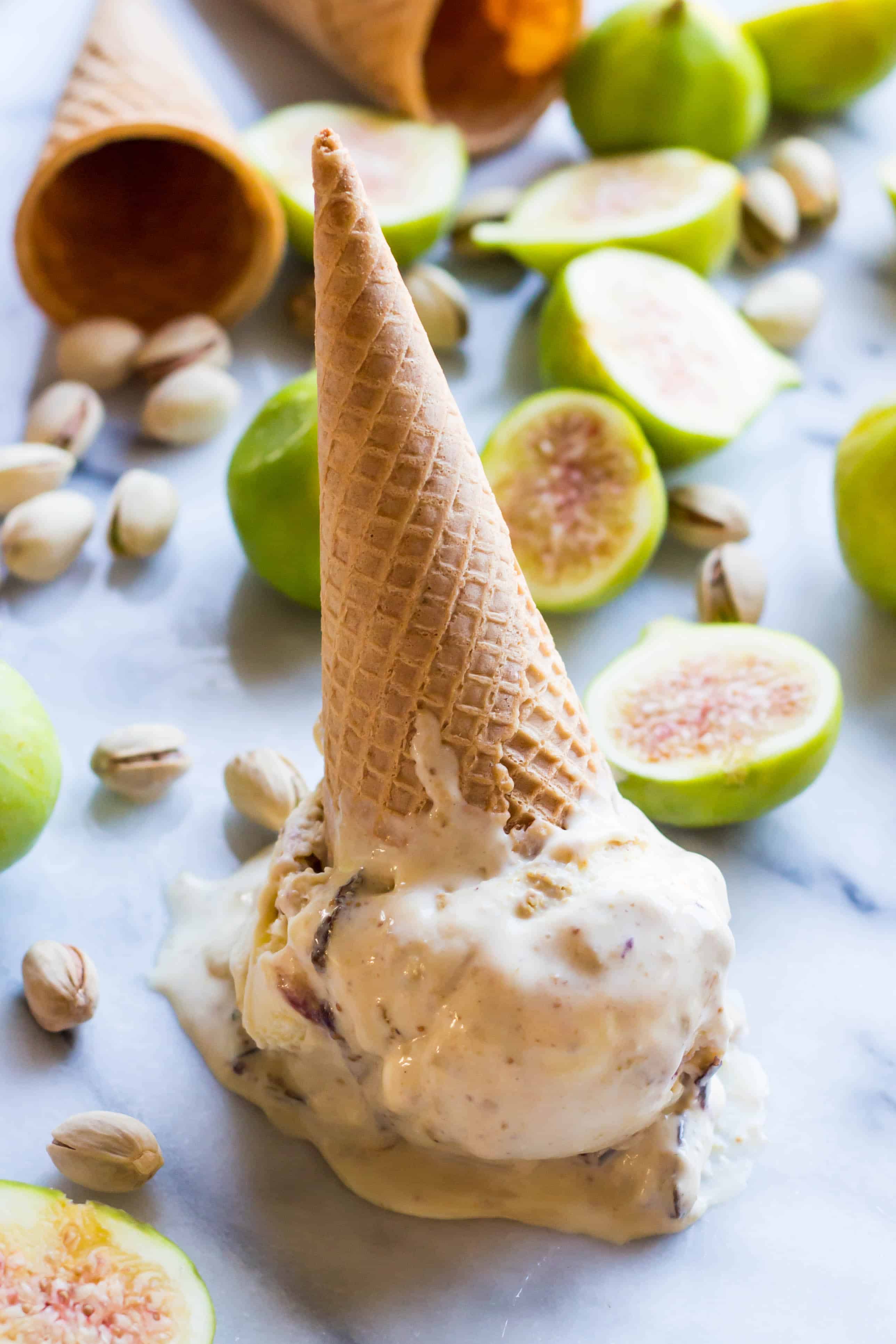 A sugar cone stuck on a large scoop of ricotta gelato of a marble counter surrounded by pistachios and fresh figs.