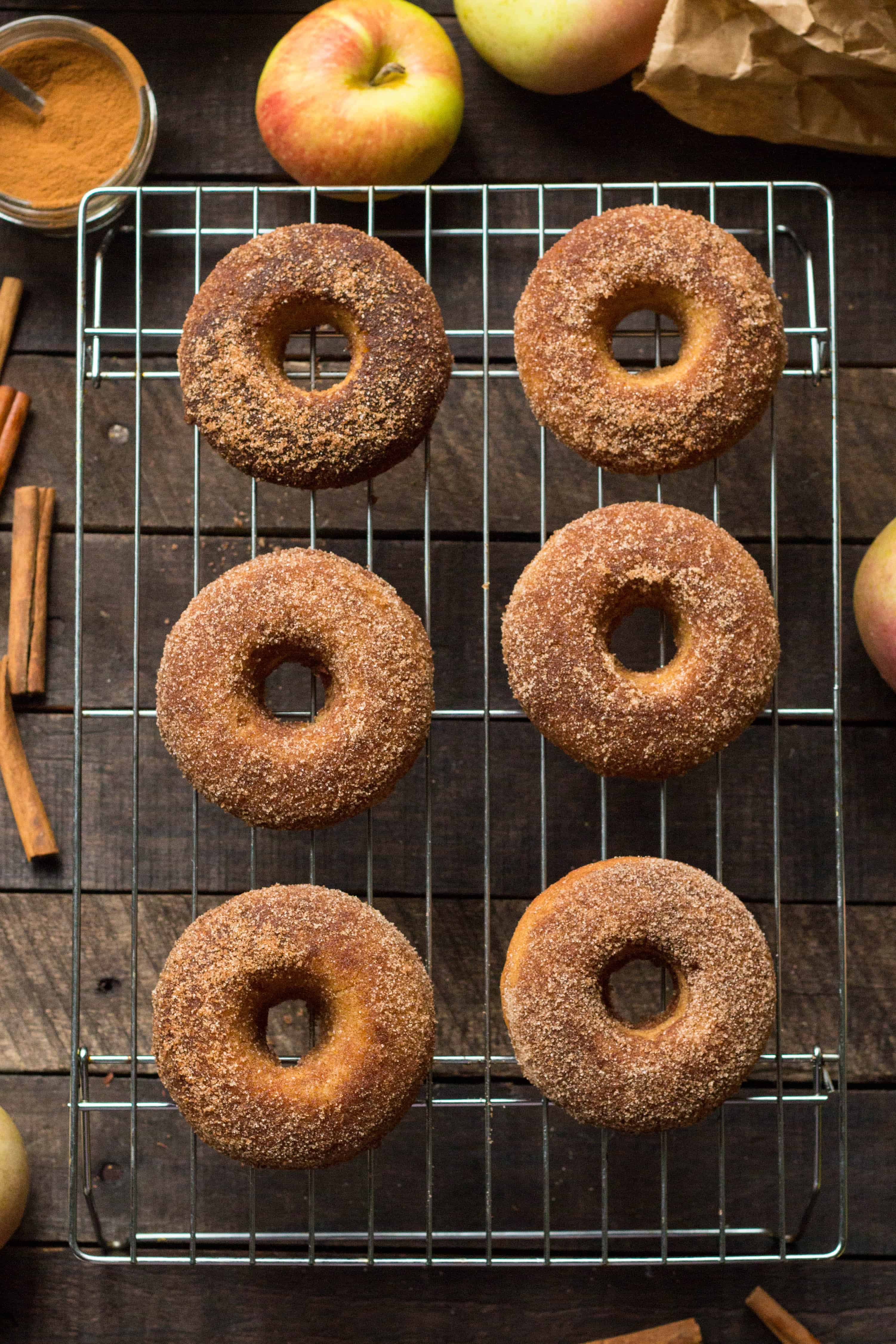 baked apple cider doughnuts cooling on a wire rack on a dark wooden table with apples and spices in the background