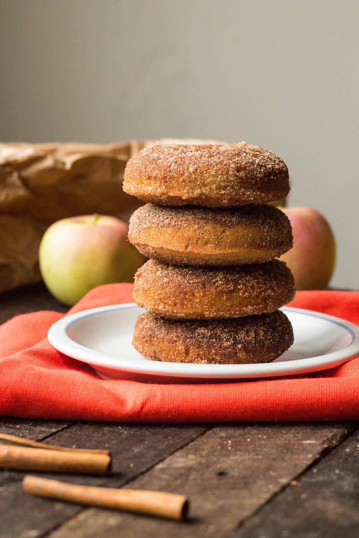 stack of baked apple cider doughnuts on a light gray plate with a red-orange napkin, an apple in the background and a cinnamon stick in the foreground