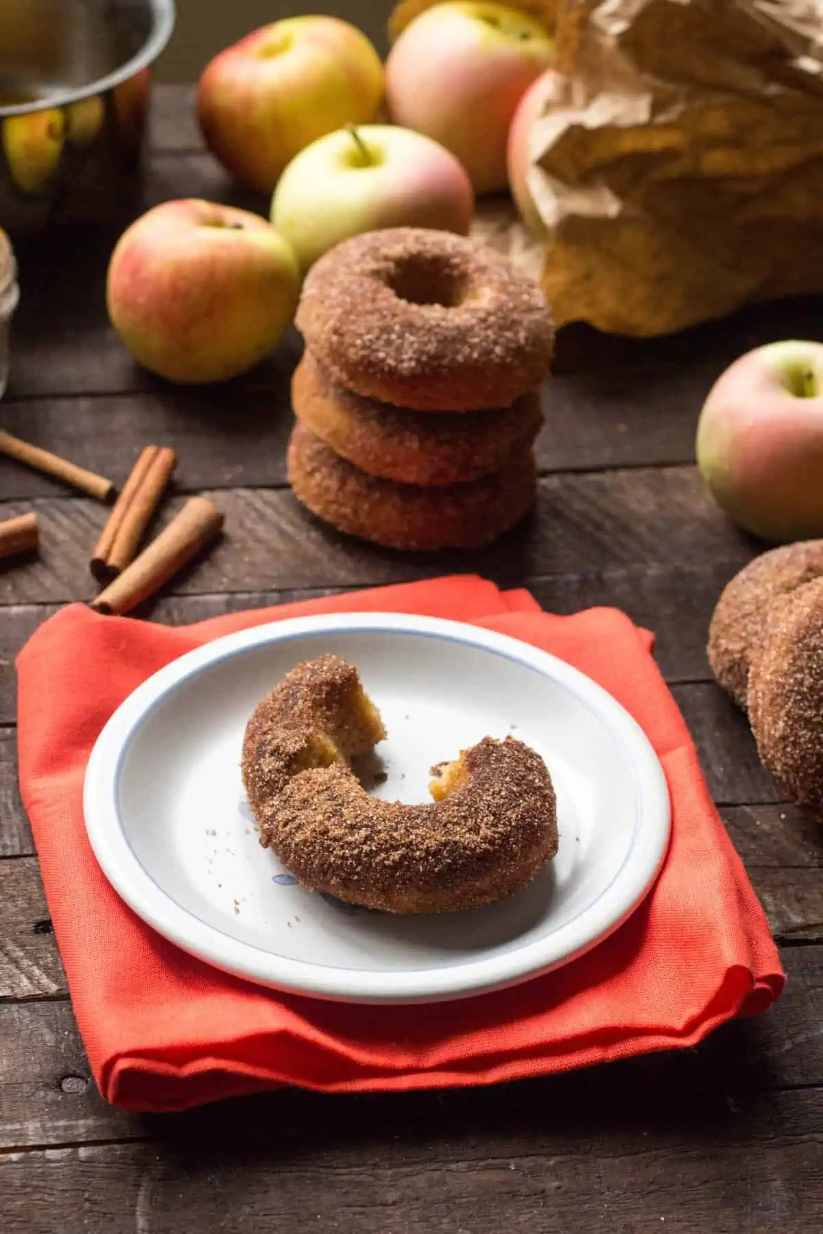 A wood table with doughnuts, apples, and cinnamon sticks around a doughnut on a small white plate.