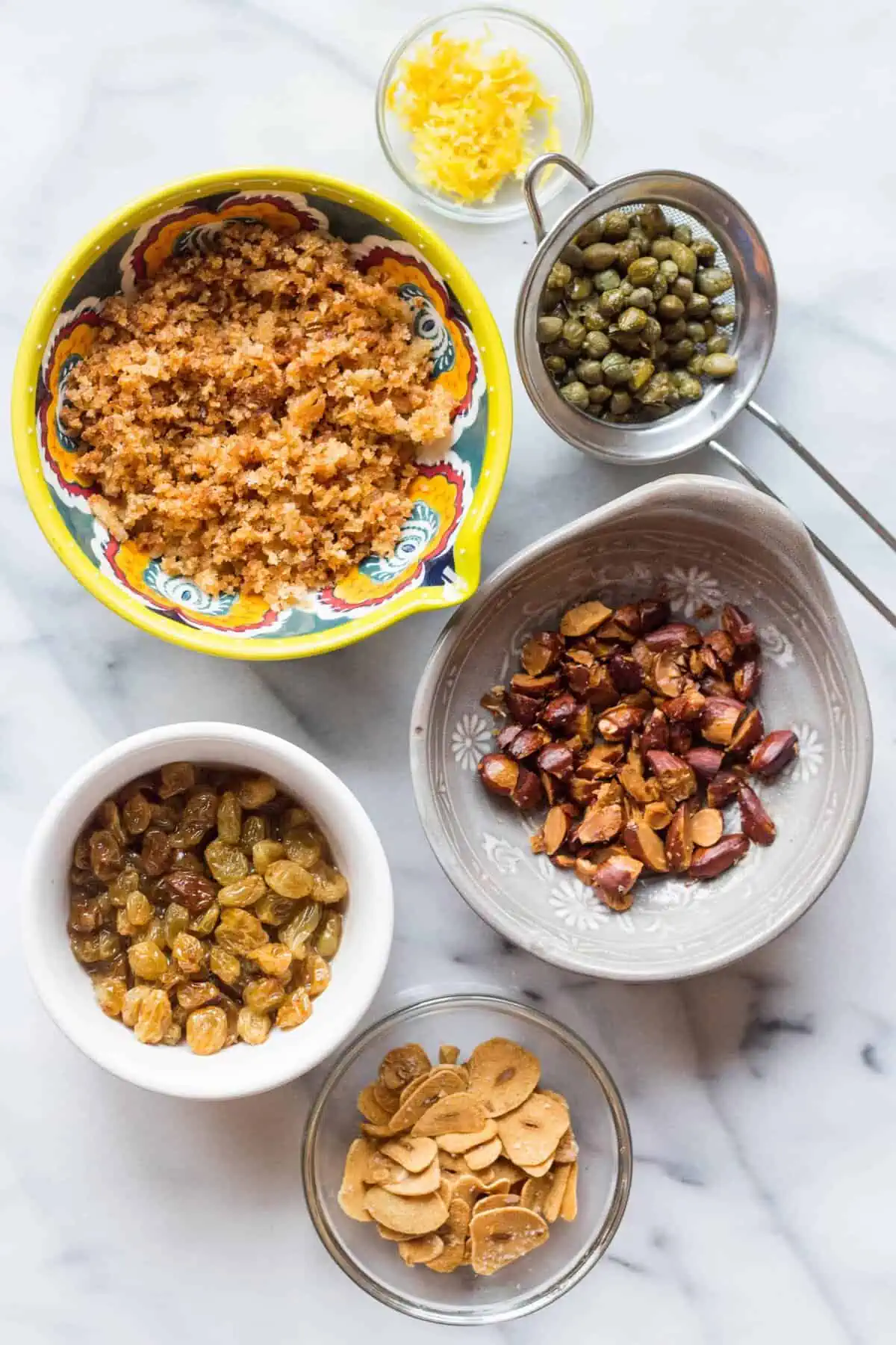 Several bowls on a marble counter filled with golden raisins, capers, toasted almonds, and capers.