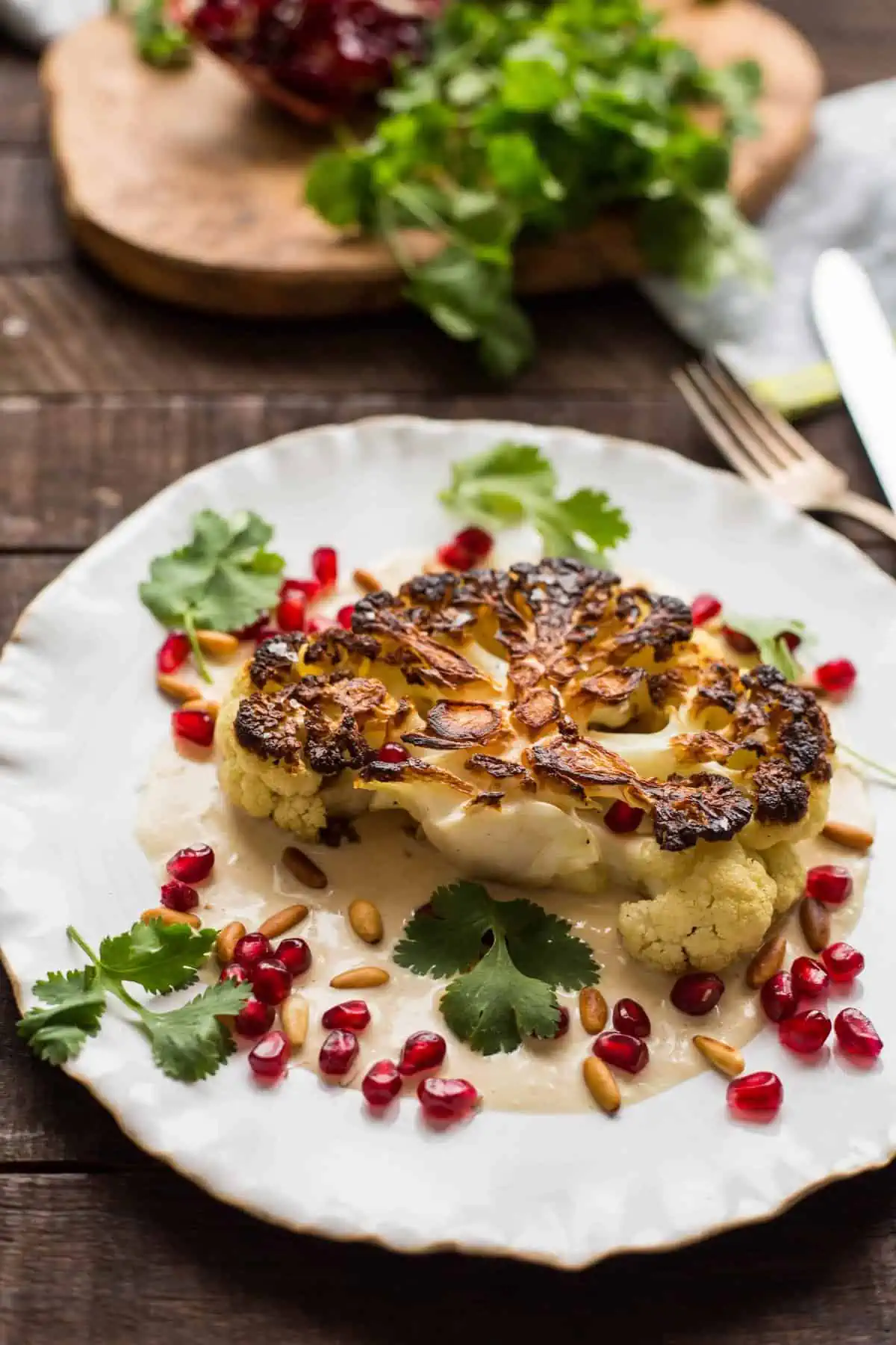A cauliflower steak surrounded by tahini, cilantro, and pomegranate arils on a white dinner plate.