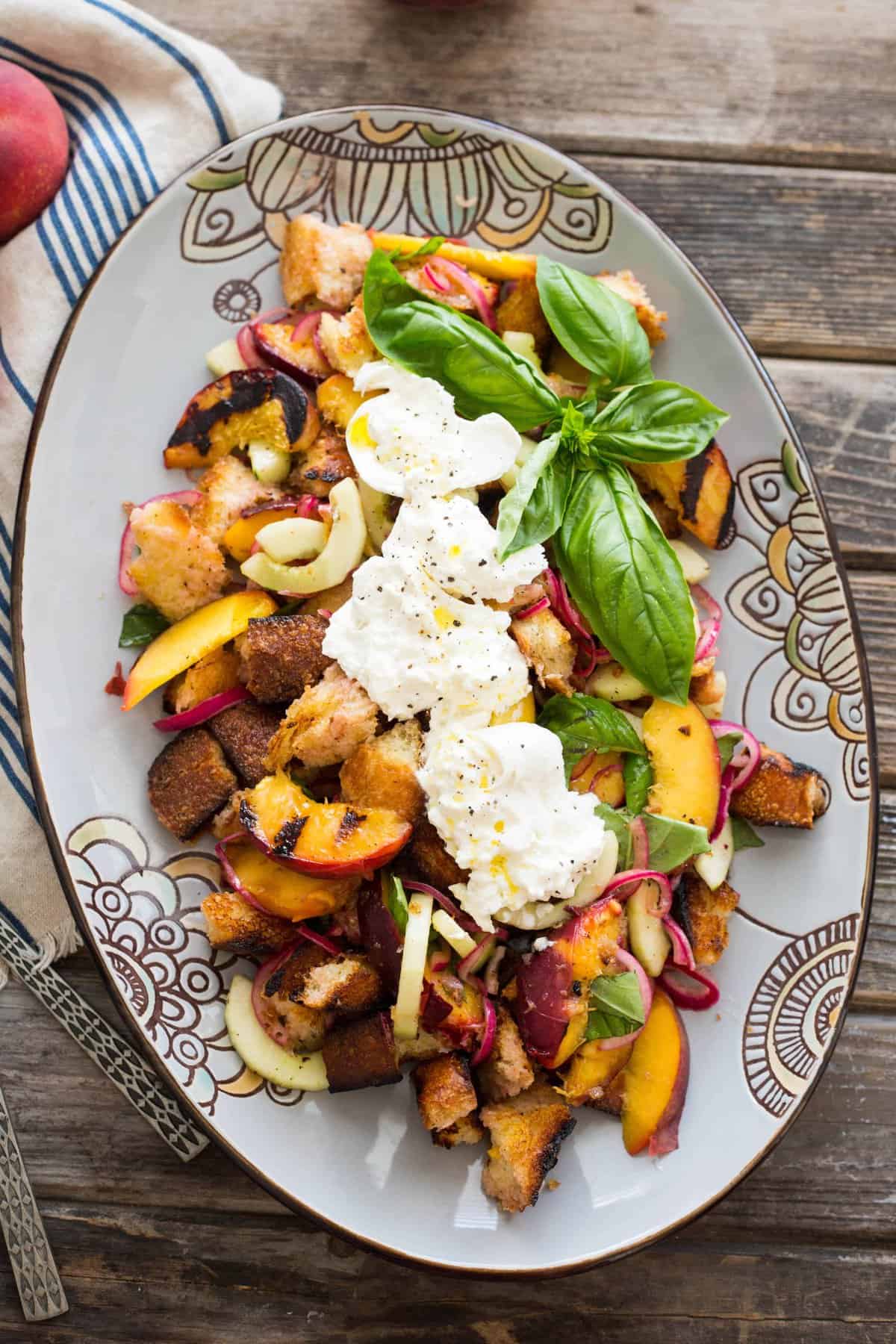 Oval serving platter of peach panzanella toped with basil and burrata.
