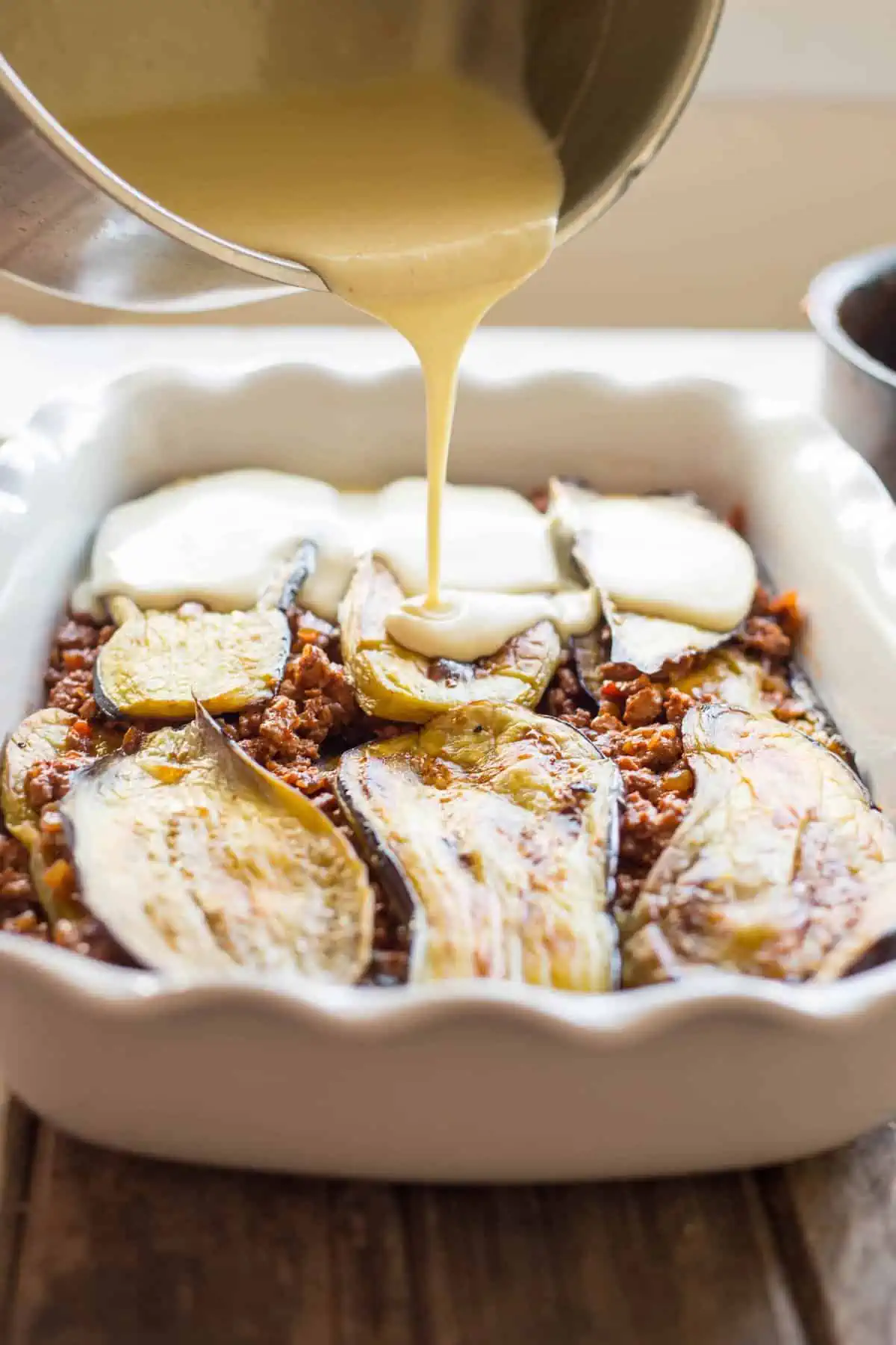 A silver pot pouring sauce overtop of layered eggplant and ground turkey in a baking dish.