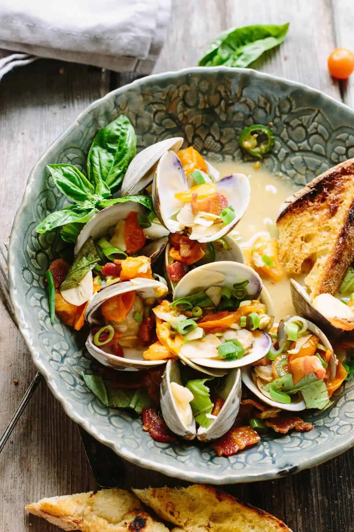 A serving bowl with tomato and jalapeno topped clams next to sliced and toasted baguette.