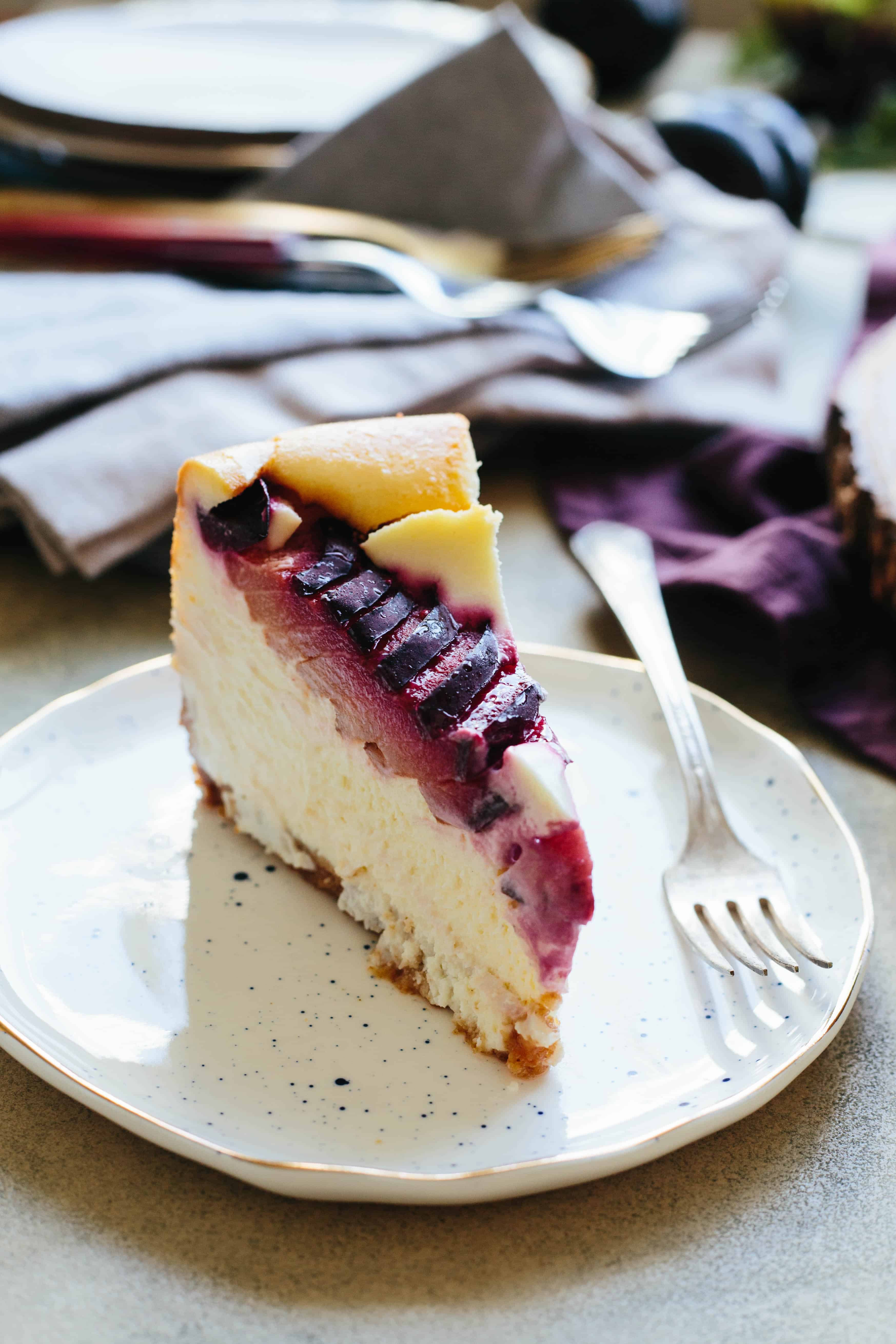 A thin slice of plum ricotta cheesecake on a speckled white dessert plate.