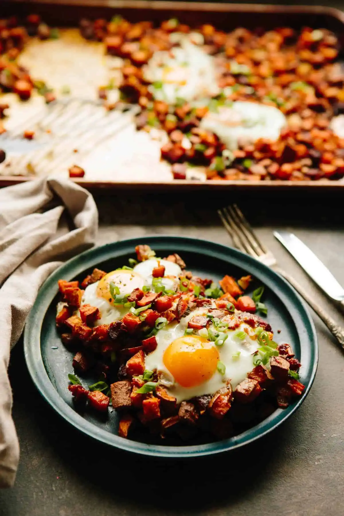 Angled shot of a dinner plate filled with sweet potato chorizo hash and runny eggs next to a baking sheet of more hash.