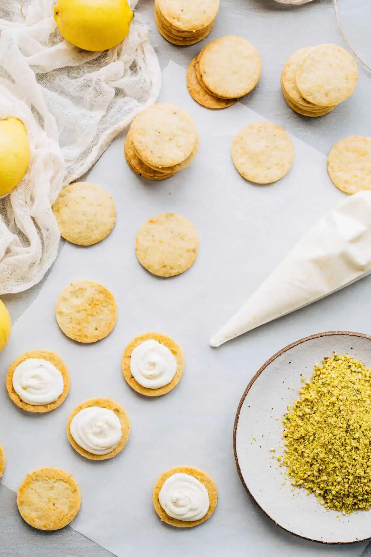A pastry bag of filling and lemon pistachio cookies on a table, half of them with filling.