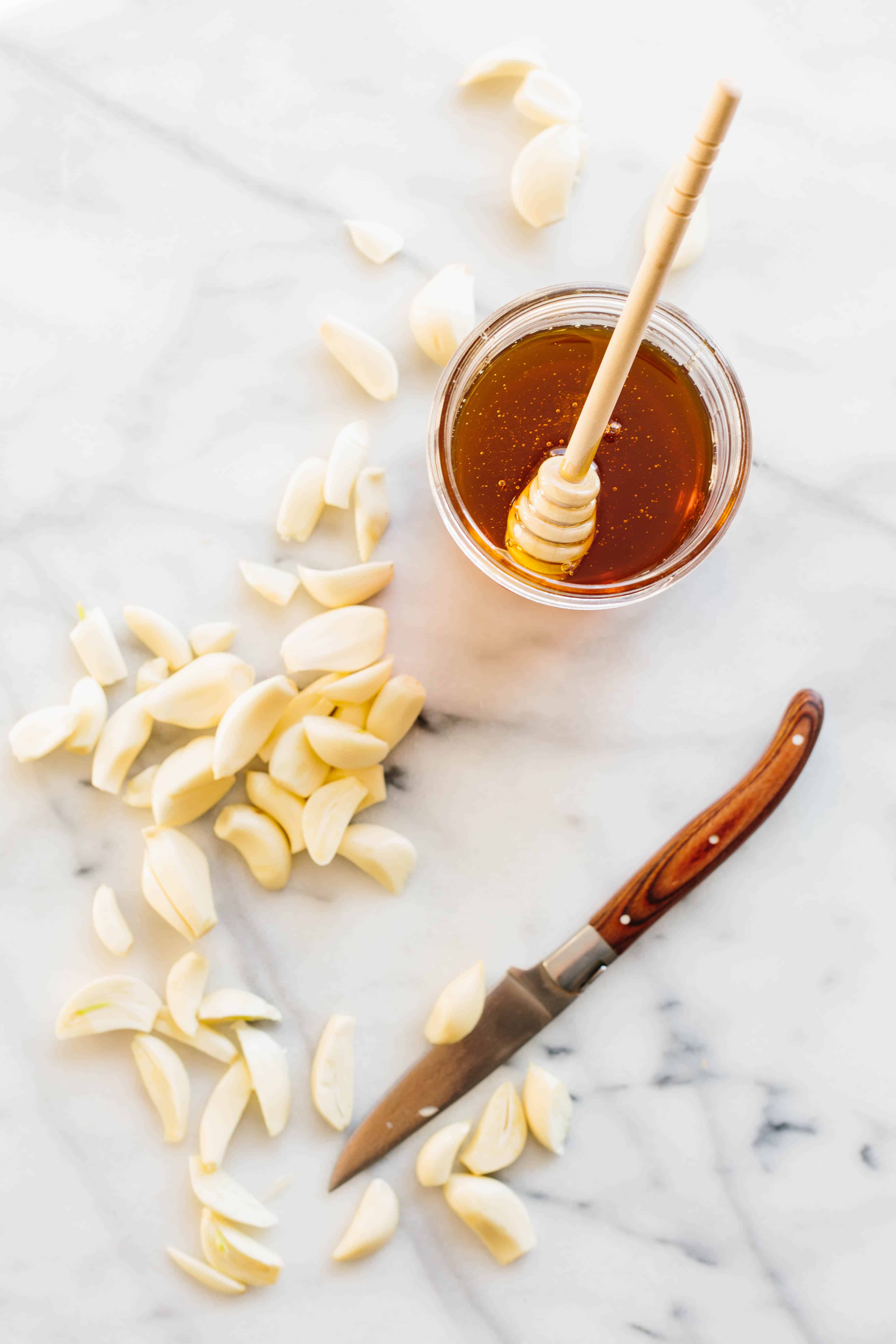 A jar of honey, a knife, and many peeled garlic cloves on a marble counter.