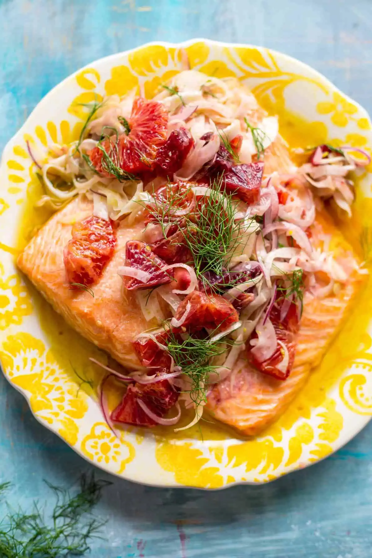 Top view of a yellow plate with a salmon fillet covered in fresh herbs and sliced citrus.