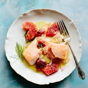 Top view of a white dinner plate with a fork, flaked salmon fillet, and sliced citrus.