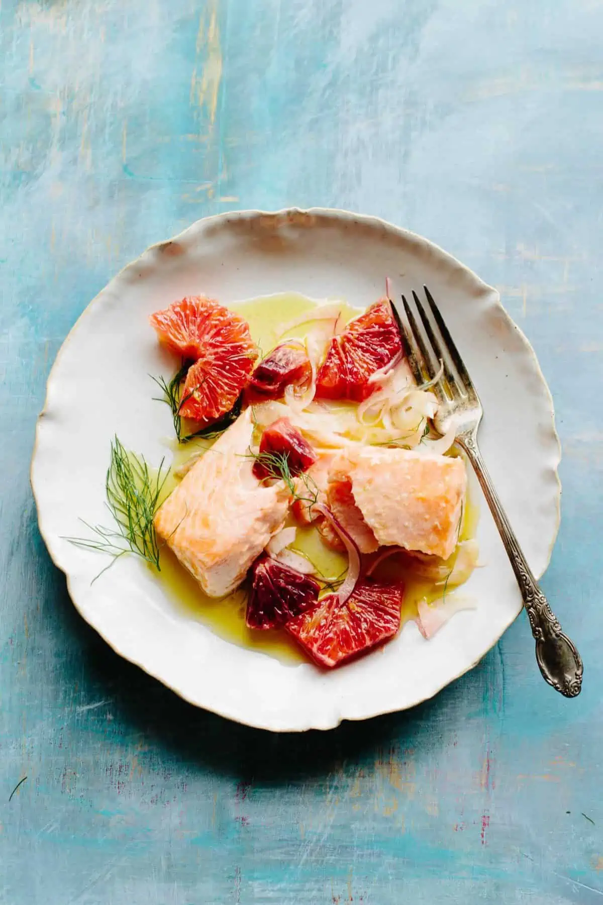 Top view of a white dinner plate with a fork, flaked salmon fillet, and sliced citrus.