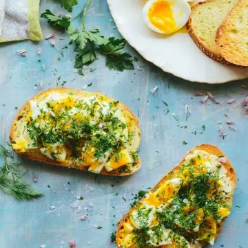 Two slices of golden toast with smashed eggs and herbs on a blue board.