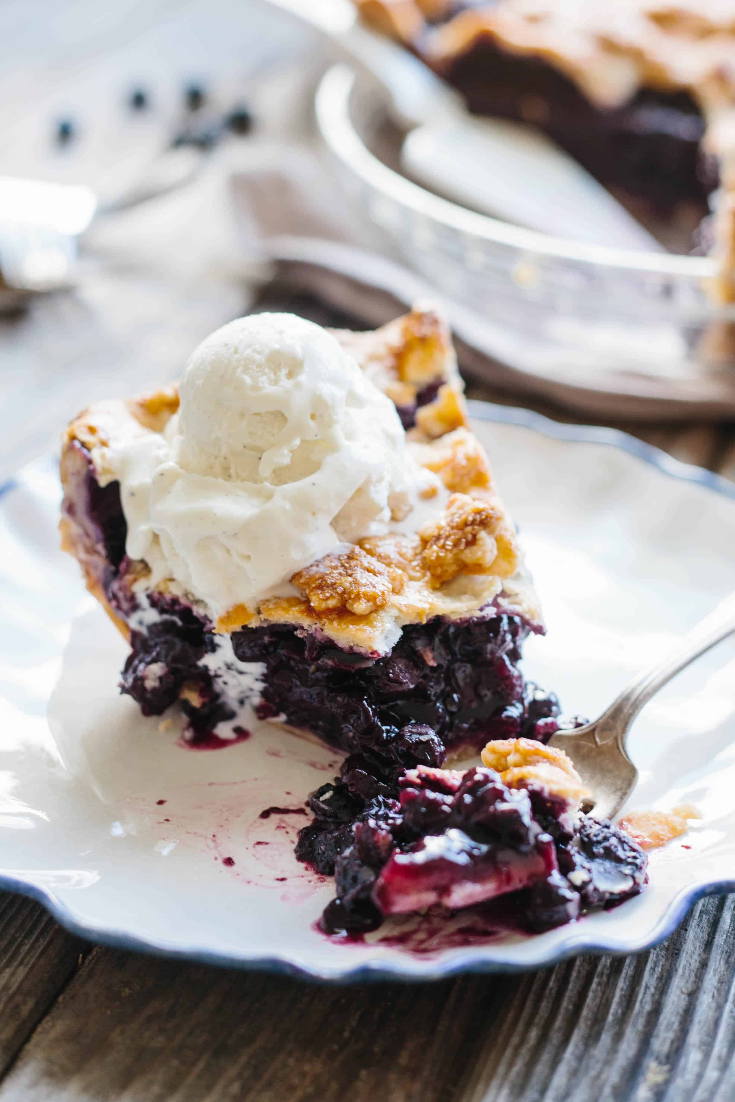 Close up of a fork grabbing a bit from a slice of blueberry pie with ice cream melting over the top.