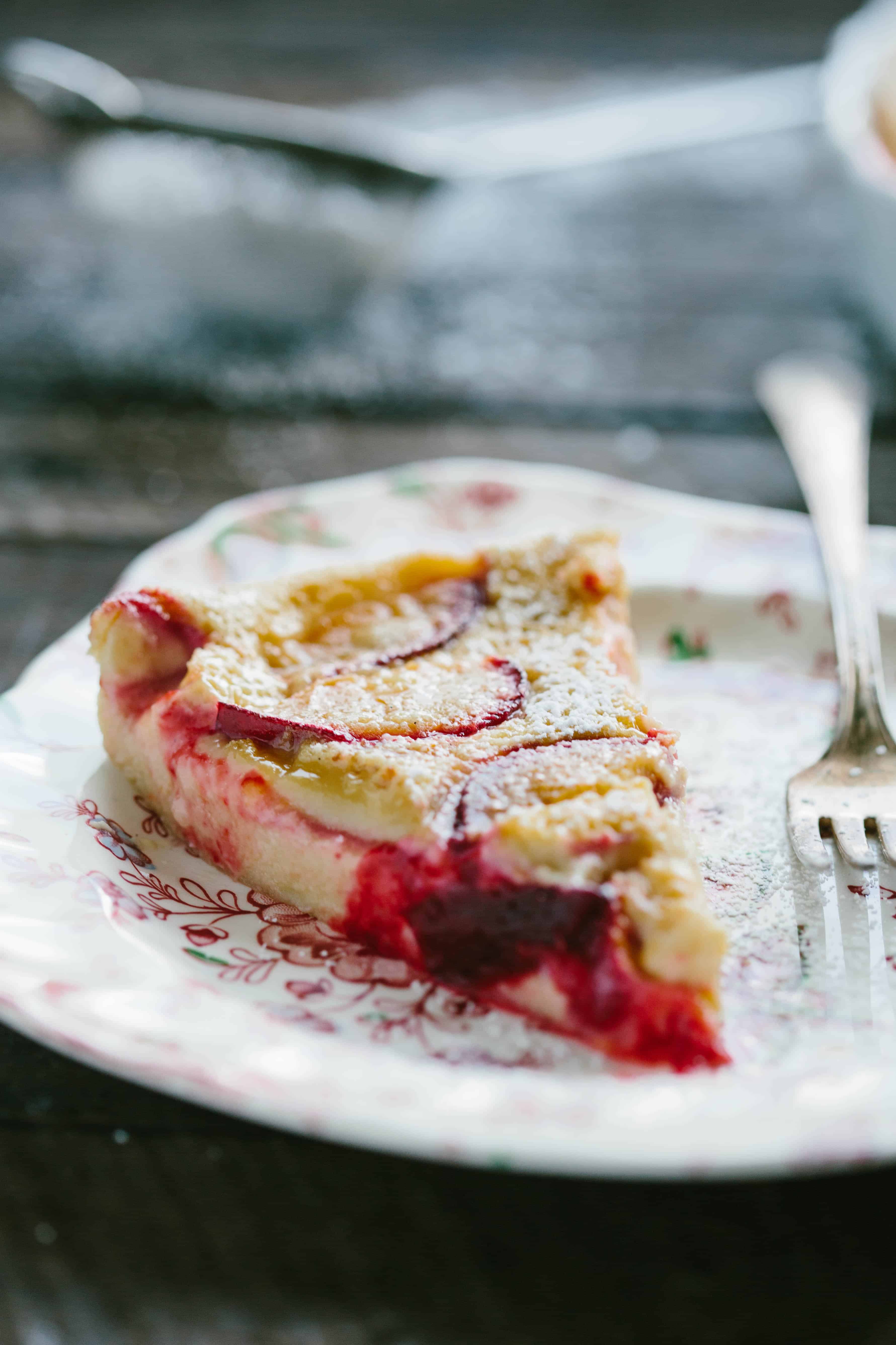Close up of a triangle slice of plum clafoutis on a flowered plate with a fork.