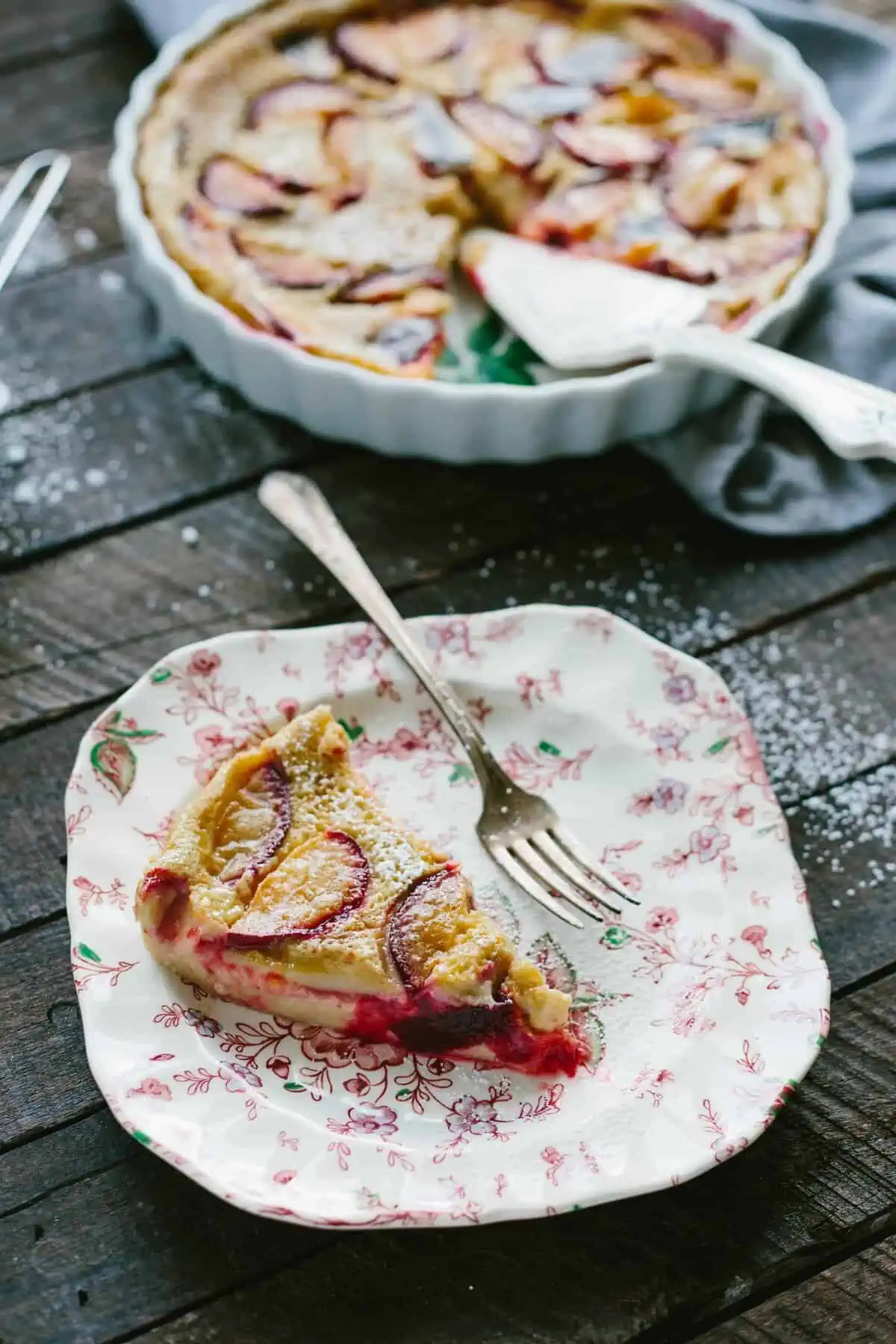 Vertical shot of a slice of plum clafoutis on a plate next to the rest of the dish in a tart pan.