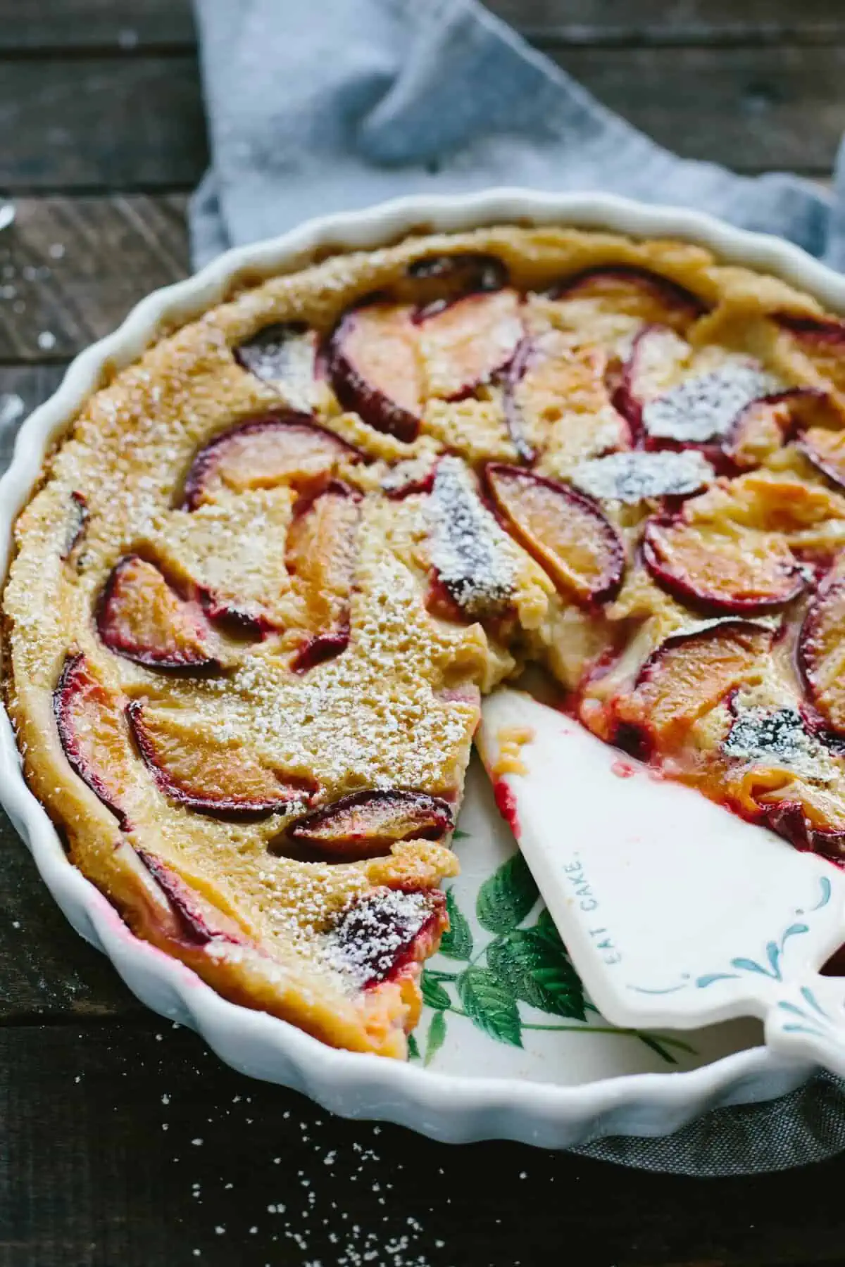Top down view of a plum clafoutis in a white baking dish with a pie server.